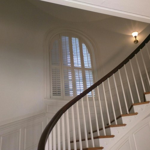 White plantation shutters decorating arched window located in spiral stairwell.